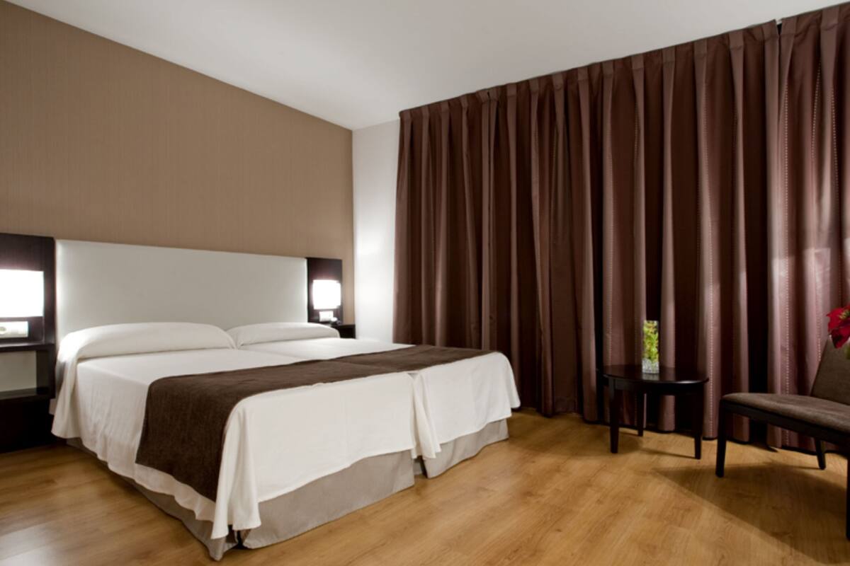 A hotel room with a large bed, a nightstand with a lamp, a small table with a chair, and a window with curtains.