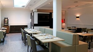 Un comedor moderno con mesas largas y asientos acolchados, una cocina al fondo y un tragaluz arriba.