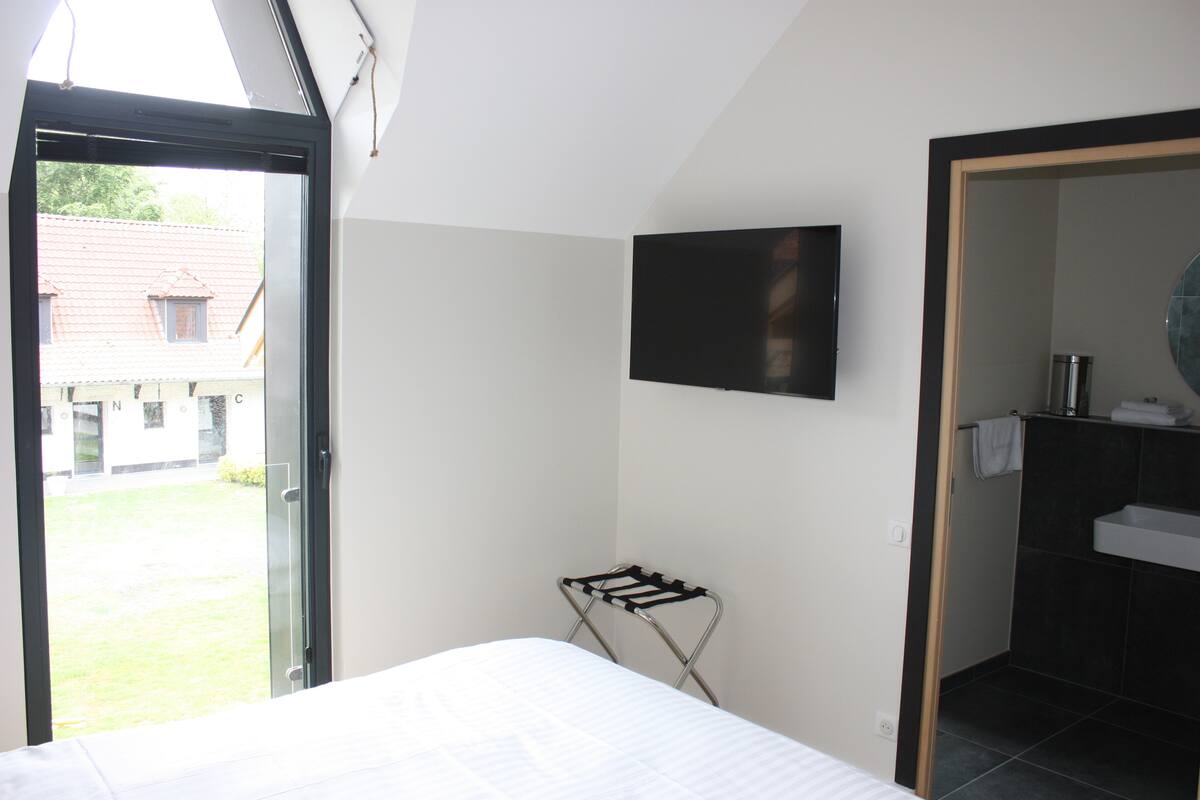 A bedroom with a bed, a black TV mounted on the wall, a window with a view of a building outside, and a bathroom with a sink and mirror.