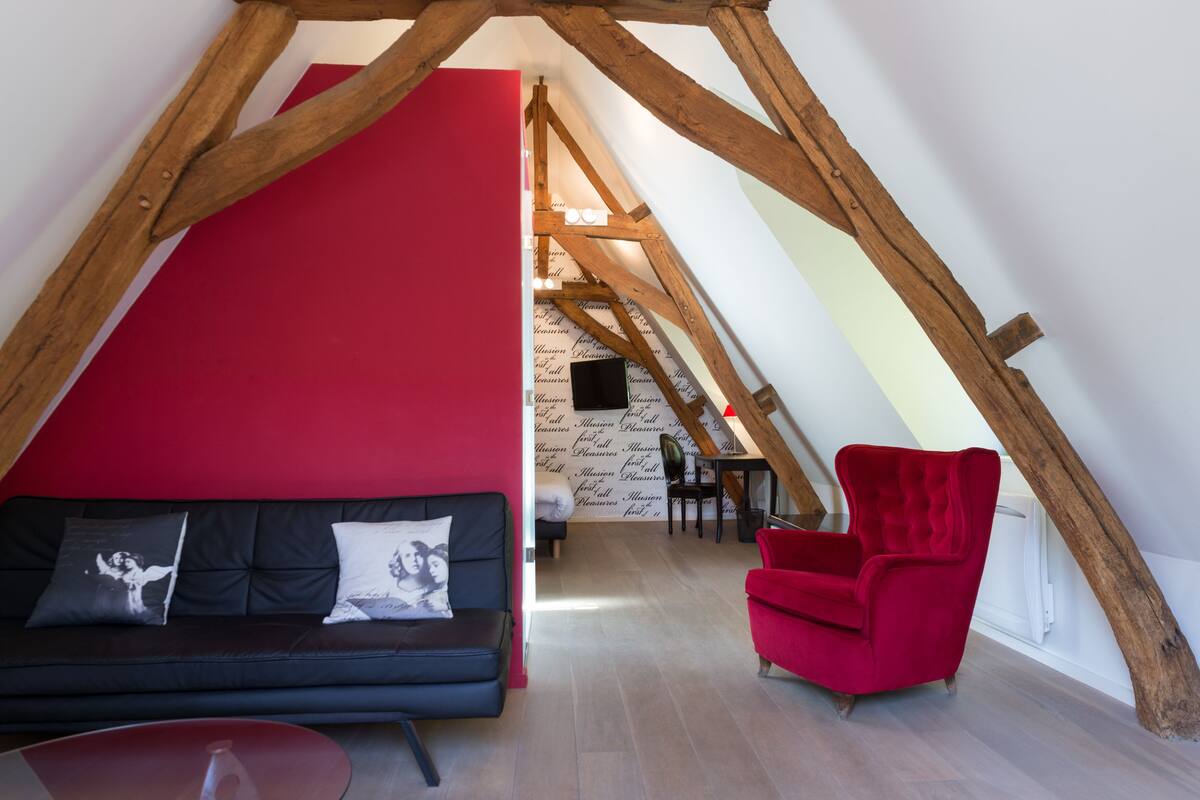 A loft-style room with a red wall, a black sofa, a red armchair, and a wooden ceiling structure.
