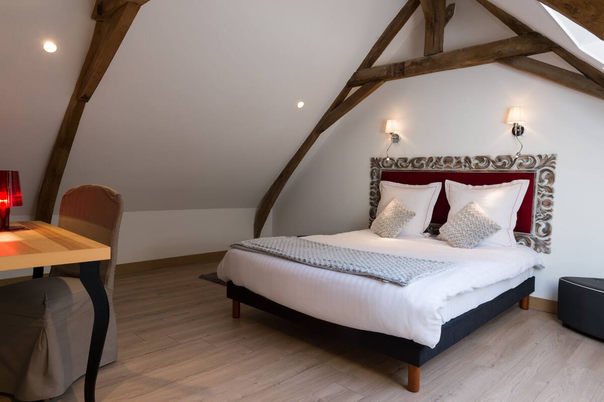 A bedroom with a wooden ceiling, a bed with white bedding, two bedside lamps, and a wooden table with a chair.