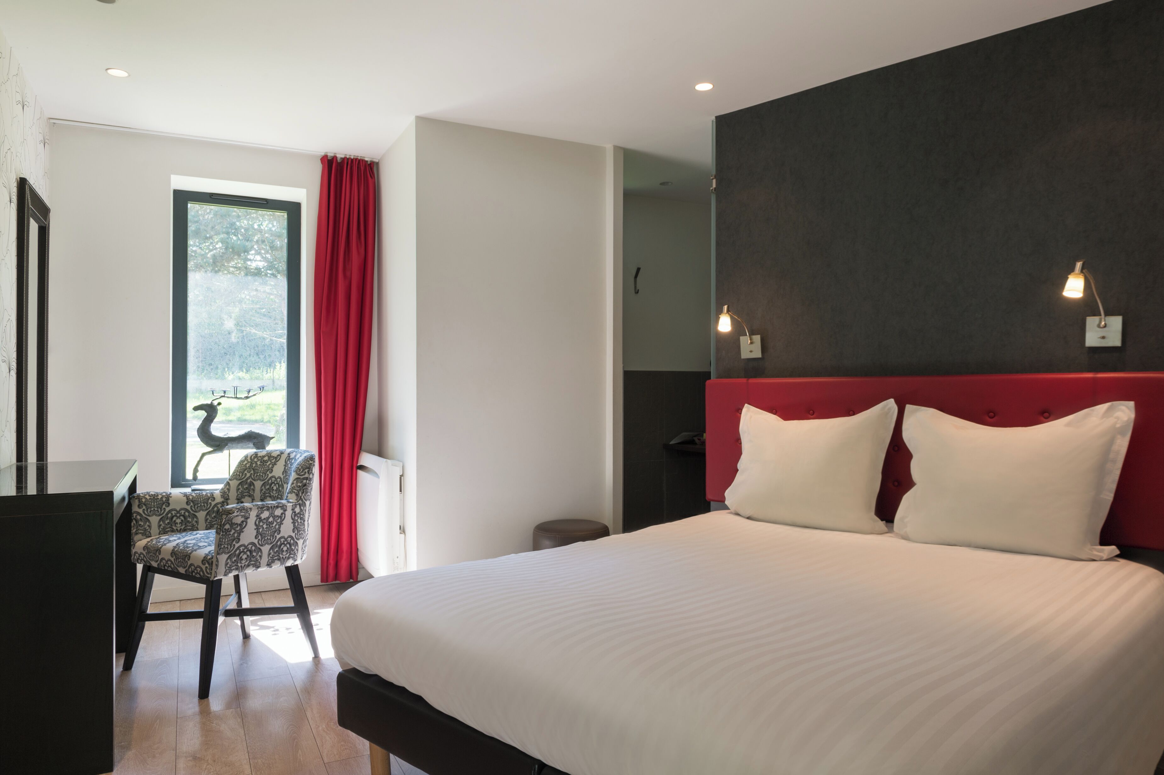 A hotel room with a bed, a desk, a chair, a window with curtains, and a view of trees.