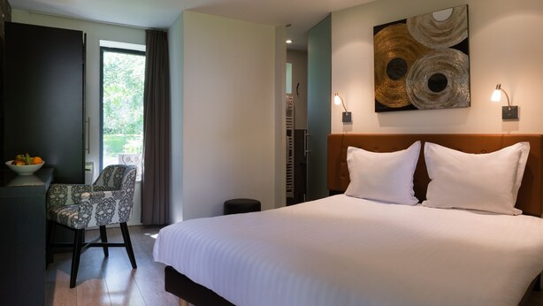 A modern hotel room with a large bed, a desk with a chair, a wall-mounted artwork, and a view through the window.
