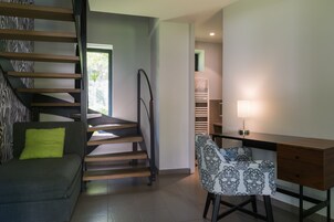 Um interior moderno com uma escada, um sofá com almofada verde, uma cadeira estampada e uma mesa de escritório com abajur.