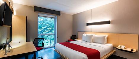A modern hotel room with a large bed, a desk, a chair, and a window with a view of trees.