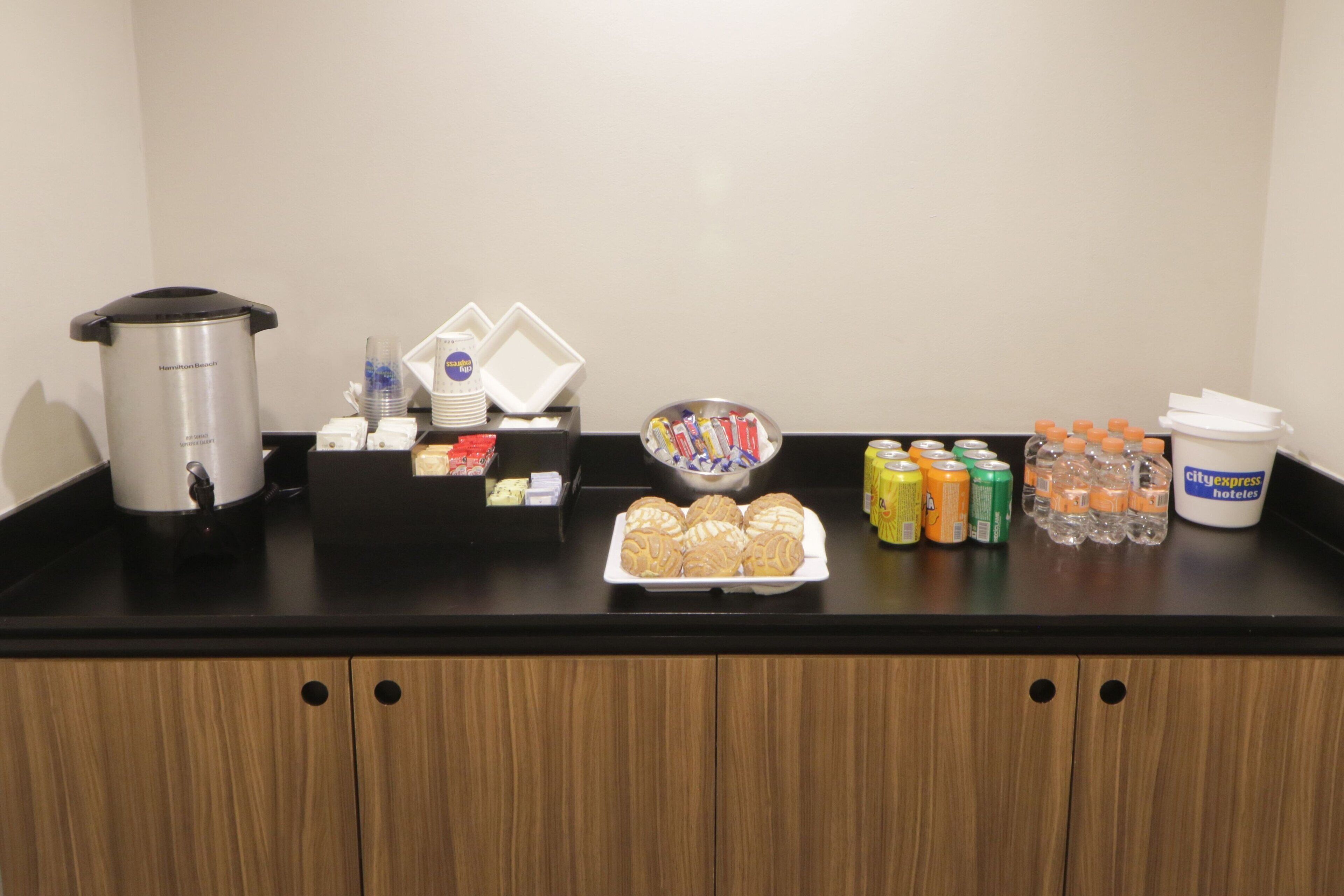 Zona de recepción de un hotel con una cafetera, bandeja de aperitivos, agua embotellada y diversas bebidas sobre el mostrador.