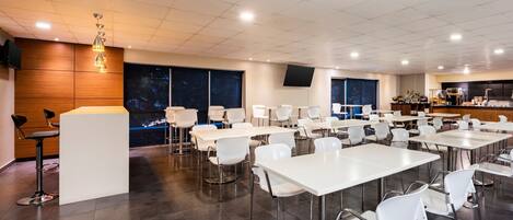 A modern dining area with white tables and chairs, a wooden accent wall, and a large window with a view of the night sky.