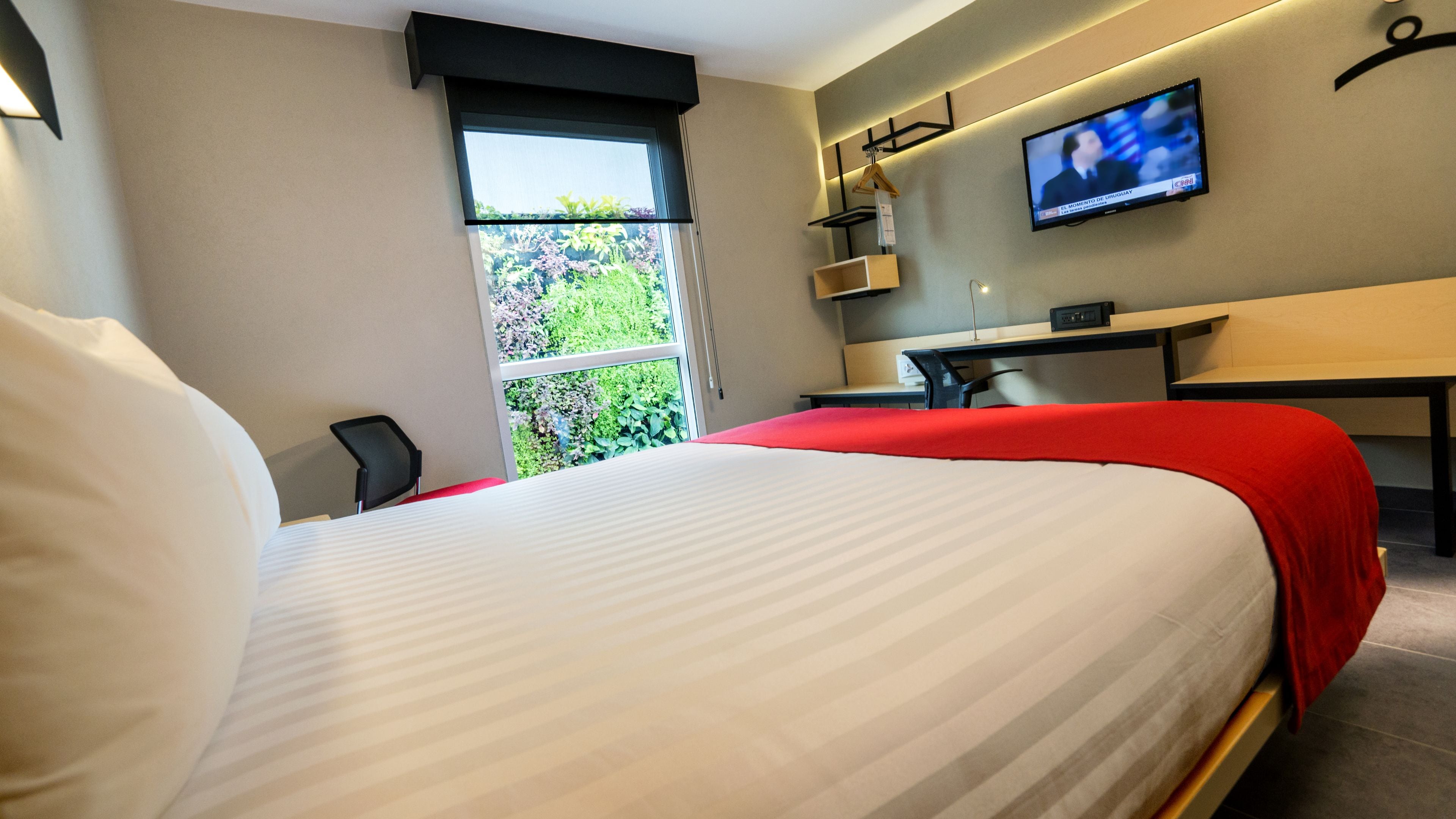A hotel room with a large bed, a desk, a chair, a TV mounted on the wall, and a window with a view of greenery.