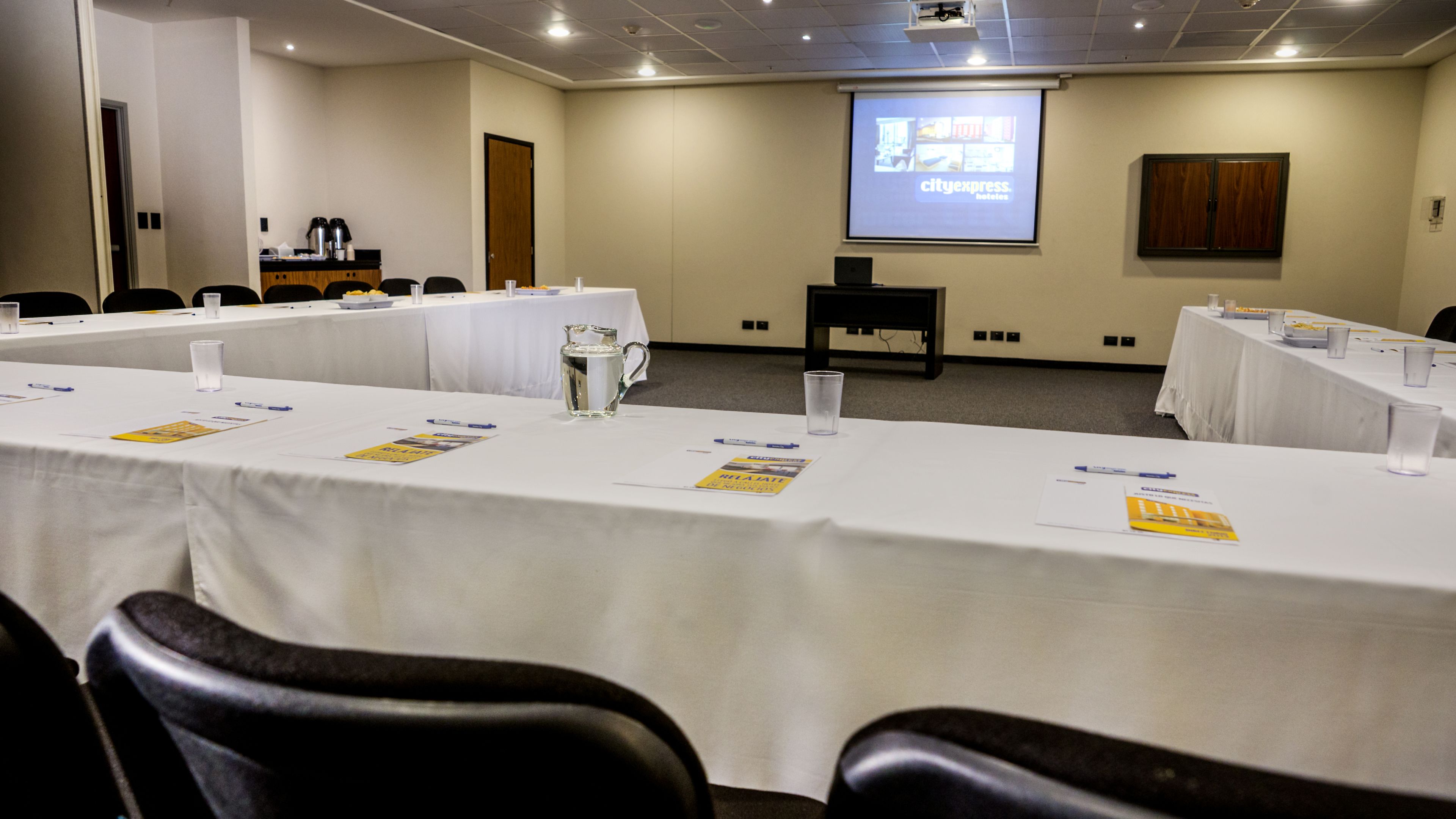 A conference room with long tables, chairs, a projector screen, and a presentation on the wall.