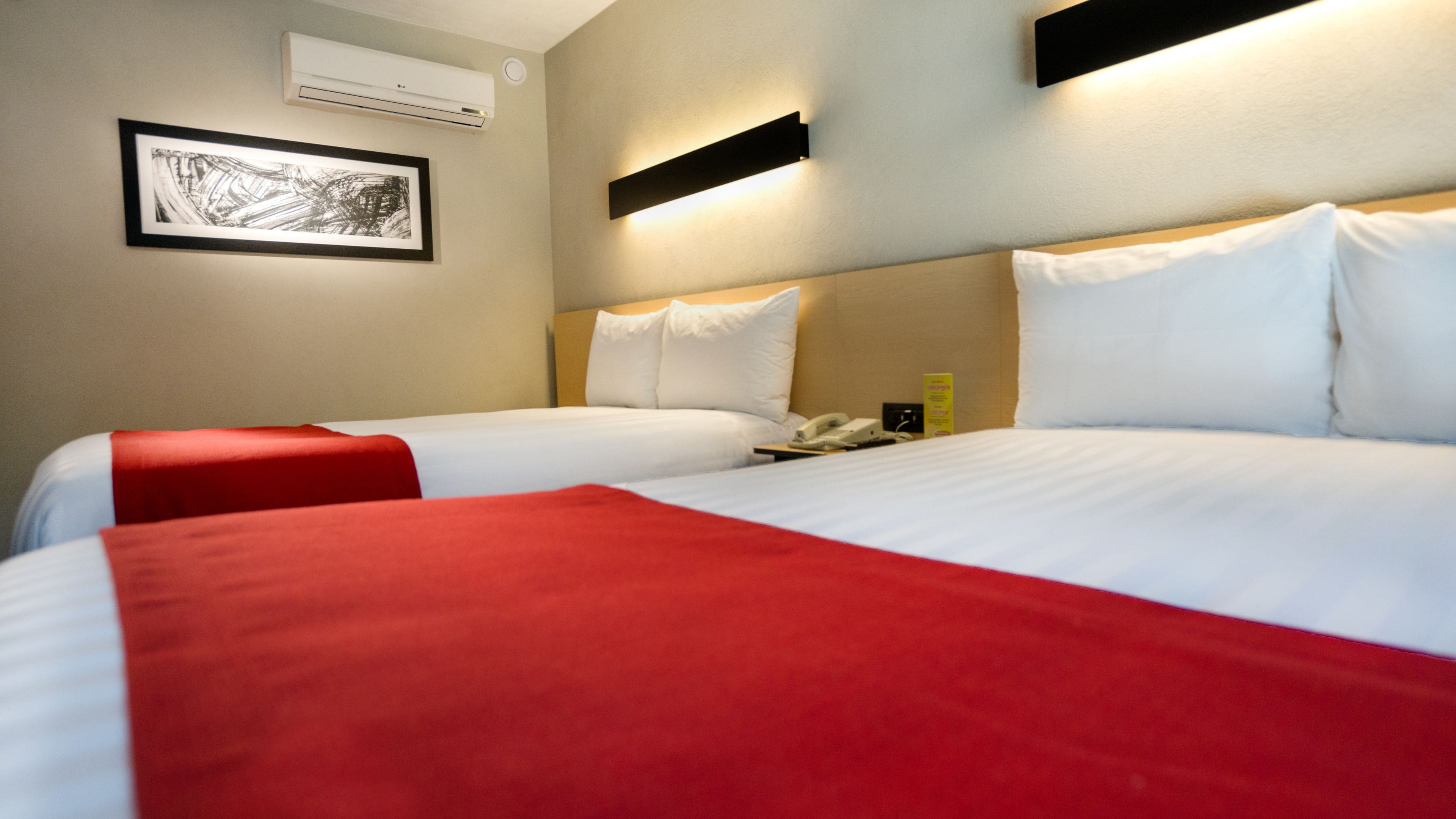 Une chambre d’hôtel avec deux lits, chacun agrémenté de draps blancs et d’oreillers décoratifs rouges. Un tableau encadré orne le mur, un téléphone se trouve sur la table de chevet, et un panneau indique les équipements disponibles.