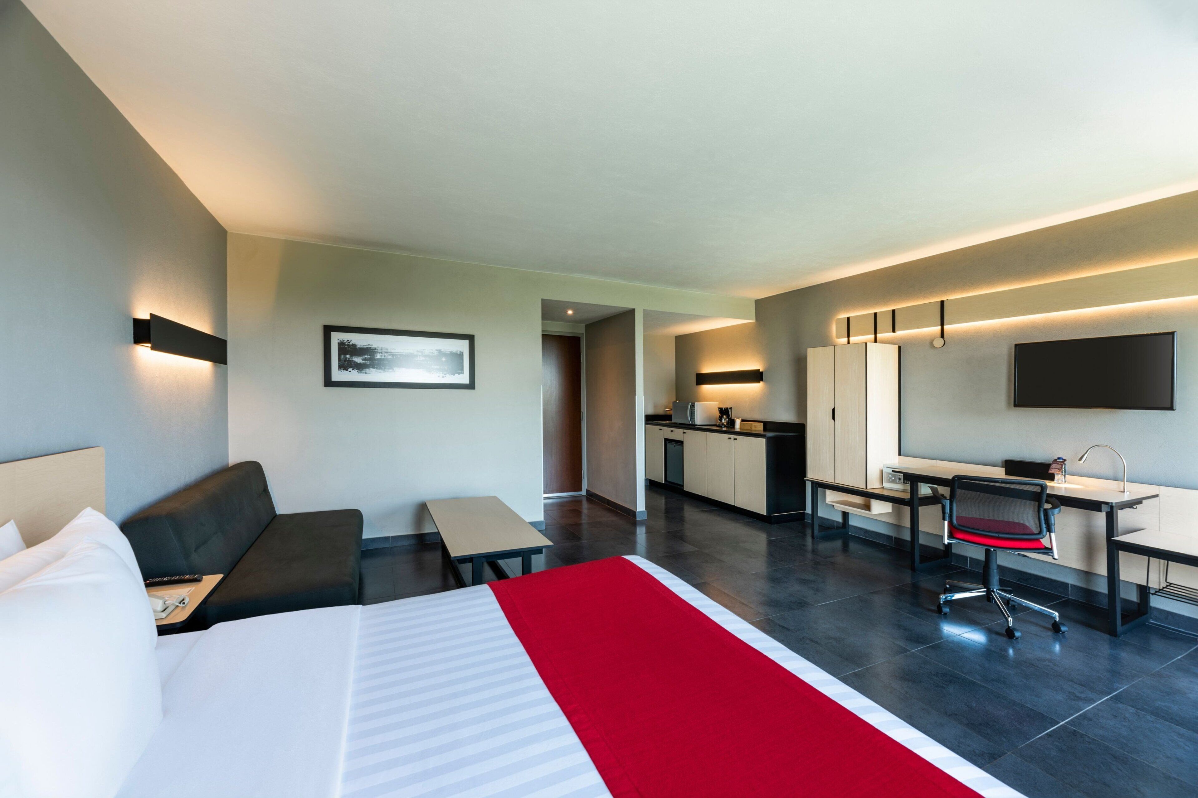 A modern hotel room with a bed, a desk, a chair, a sofa, a kitchen area, and a TV.