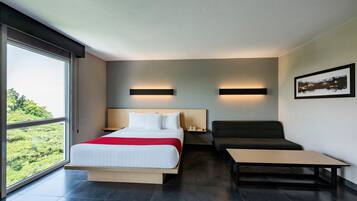 Habitación de hotel moderna con una cama grande, un sofá, una mesita y vistas al verdor a través de amplias ventanas.