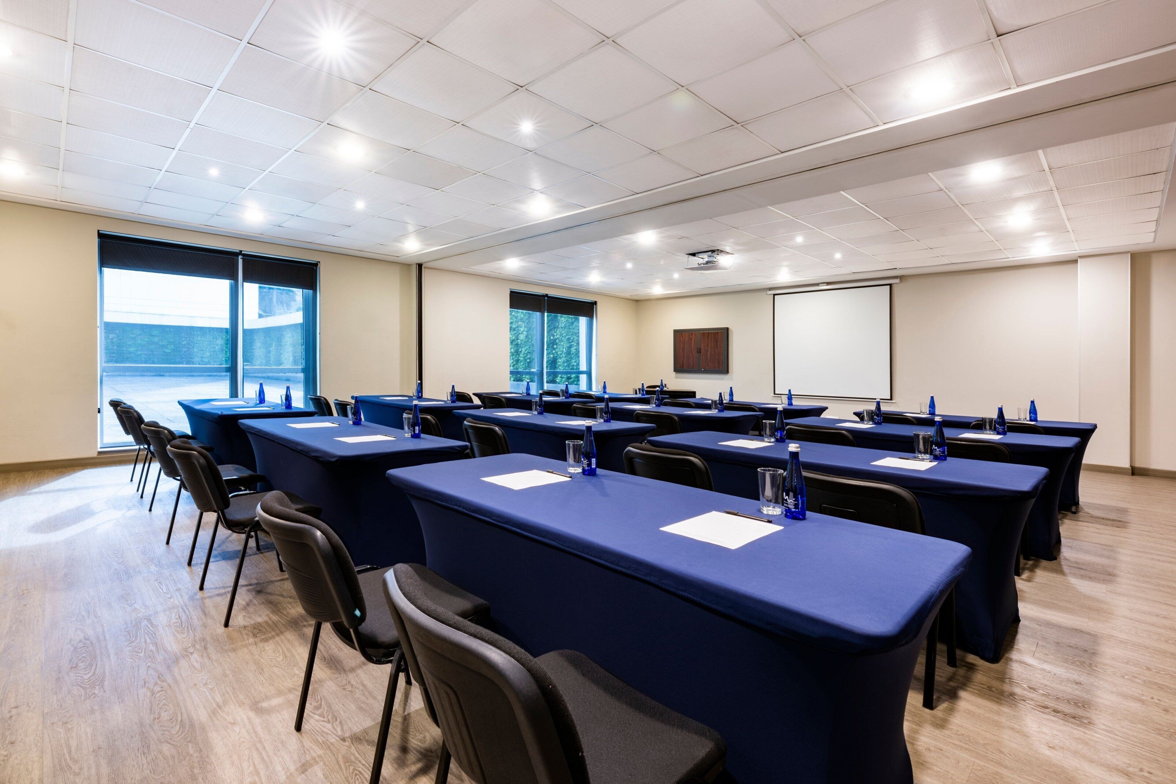Una sala de conferencias con varias mesas y sillas, ventanas amplias y una pantalla para proyector.