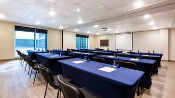 Una sala de conferencias con varias mesas y sillas, ventanas amplias y una pantalla para proyector.