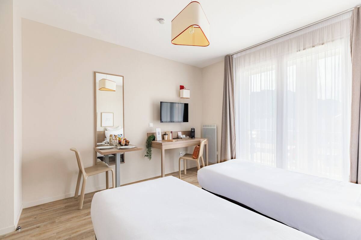 Une chambre d’hôtel avec un grand lit, un bureau avec un ordinateur, une télévision et une fenêtre avec des rideaux légers.