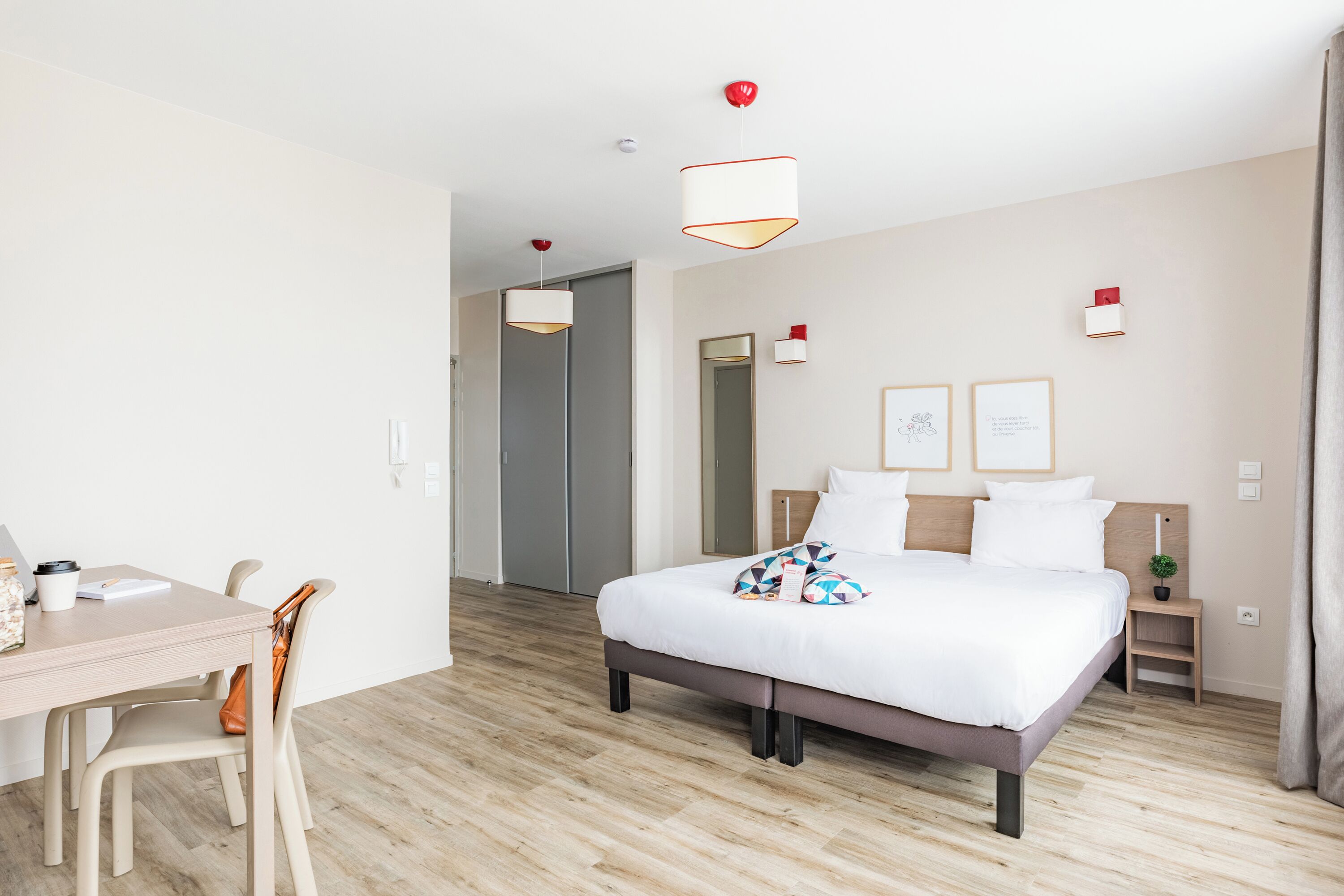 A modern hotel room with a wooden floor, a bed with white bedding, a desk with a chair, and a small bedside table with a lamp.