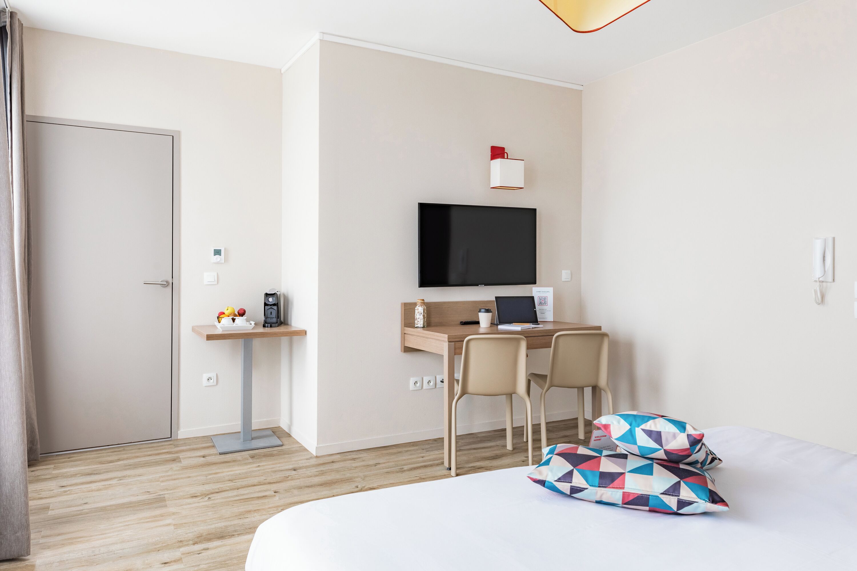A hotel room with a bed, a desk with a computer, a TV, and a small table with a cup and a fruit bowl.