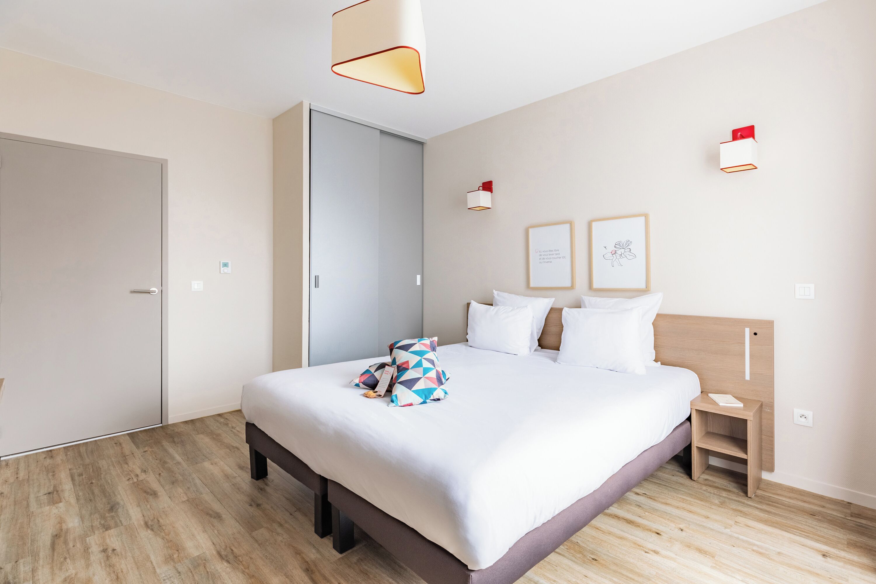 A modern hotel room with a large bed, a bedside table, a wardrobe, and two wall-mounted light fixtures.