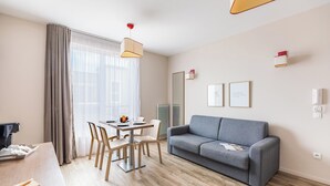 Ein modernes Hotelzimmer mit Essbereich, einem grauen Sofa und einem großen Fenster mit durchsichtigen Vorhängen.