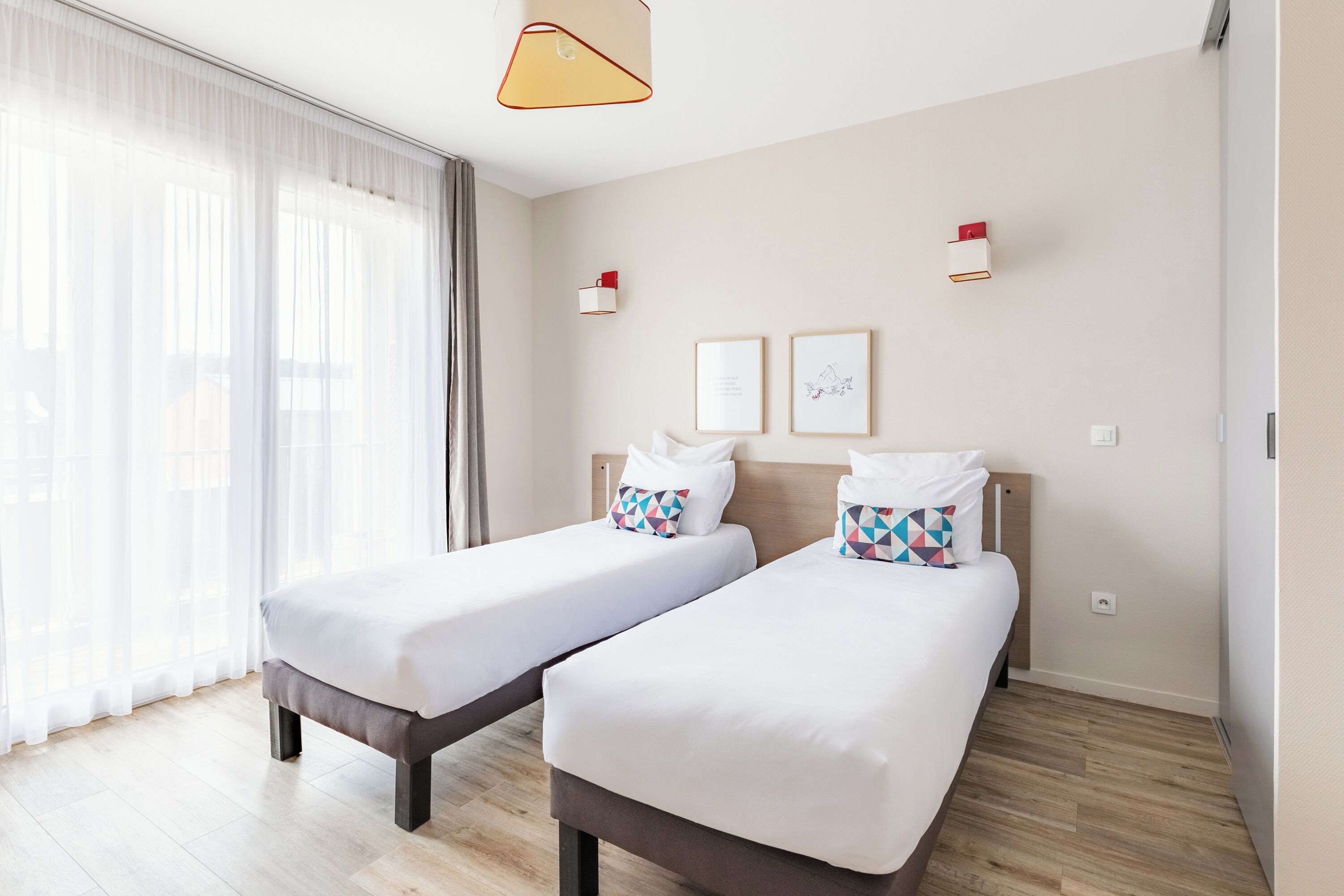 A hotel room with two single beds, a large window with sheer curtains, a wooden floor, and a modern pendant light.