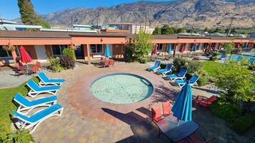 A pool area with lounge chairs and umbrellas, surrounded by buildings with a mountainous backdrop.