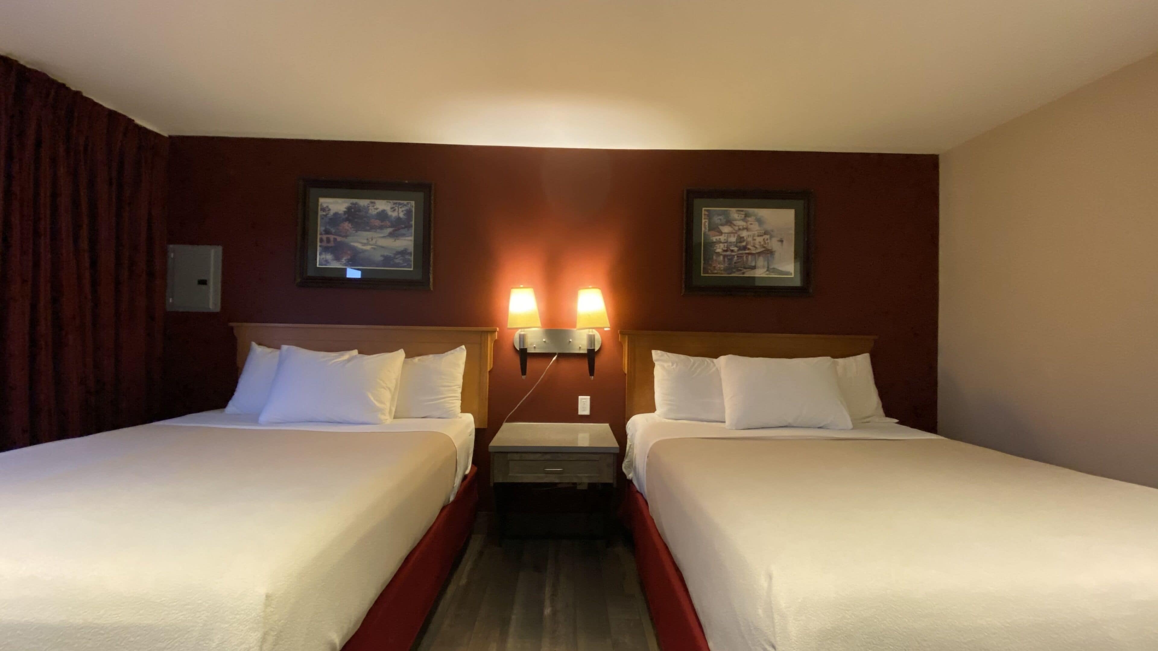 A hotel room with two beds, a wooden headboard, a small table, and two wall-mounted lamps.