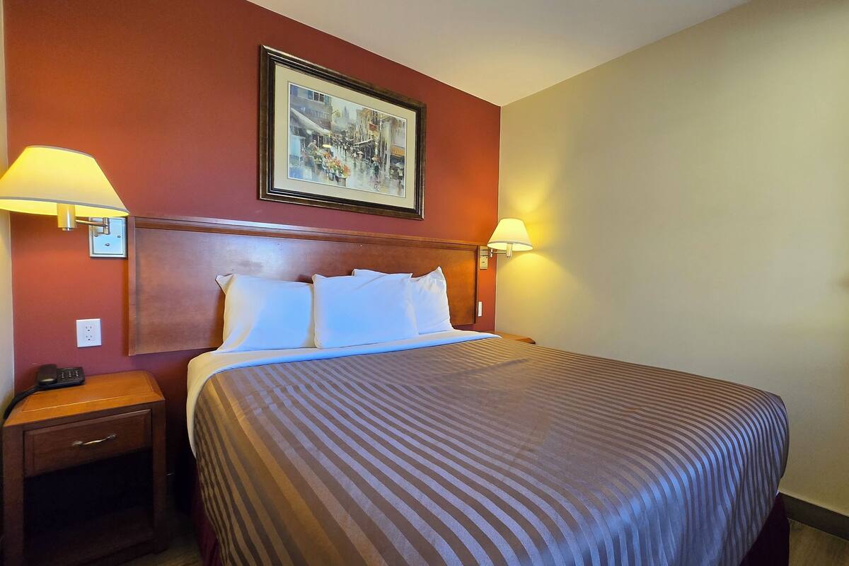 A hotel room with a bed, two bedside lamps, a nightstand with a phone, and a framed picture on the wall.