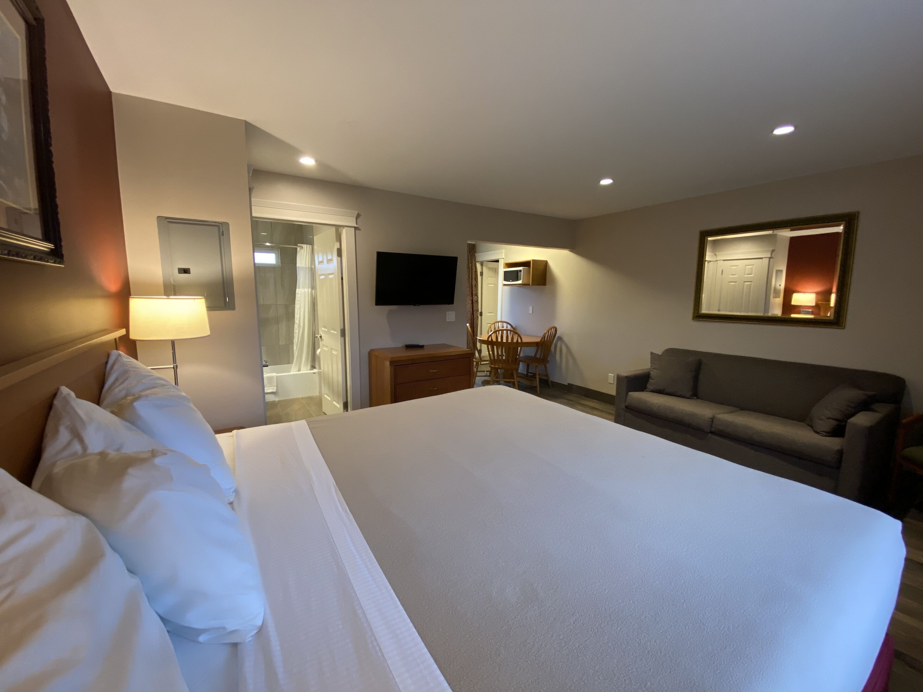 A hotel room with a large bed, a desk with a chair, a sofa, a TV, and a bathroom with a shower and toilet.