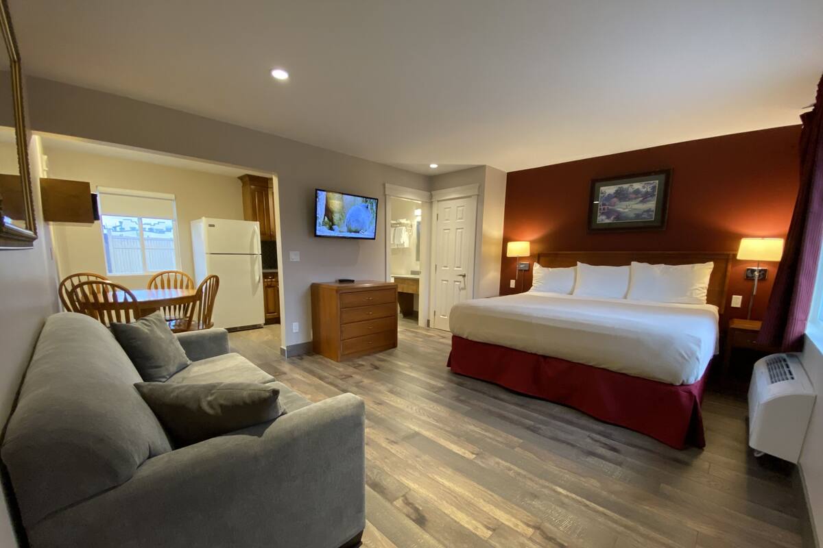 A hotel room with a bed, a sofa, a TV, a dresser, and a kitchen area visible through an open door.