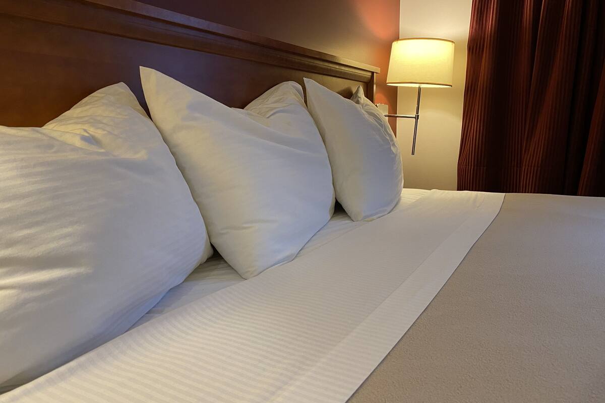 A neatly made bed with white linens and pillows, a wooden headboard, and a wall-mounted lamp.
