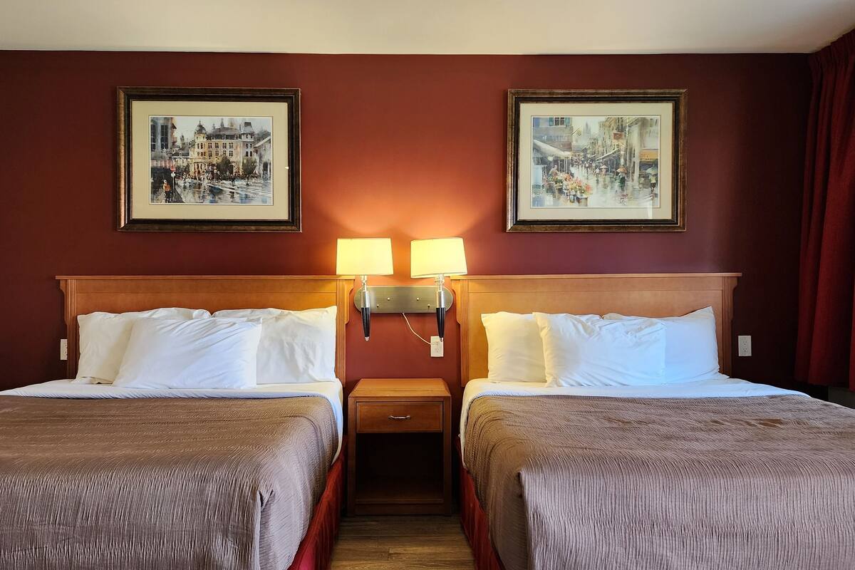 A hotel room with two beds, a wooden headboard, two wall-mounted lamps, and two framed pictures on the wall.
