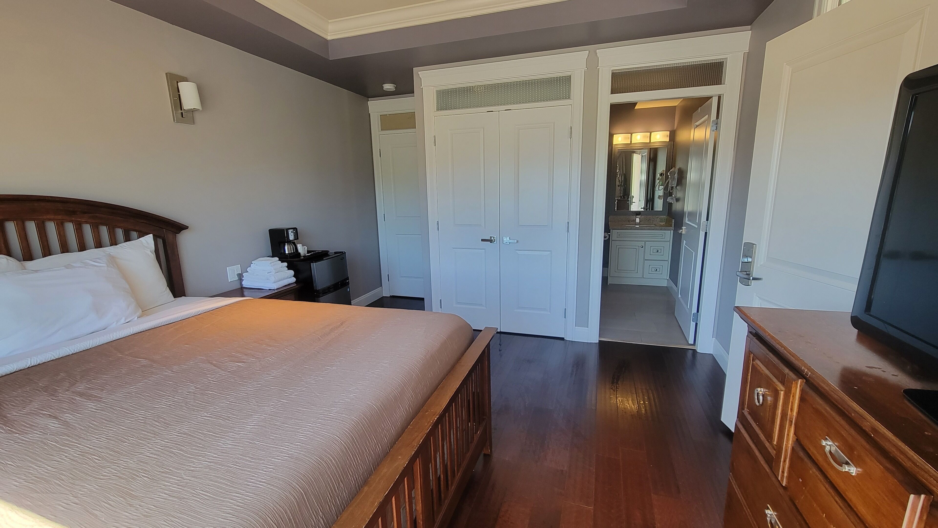 Quarto de hotel com uma cama, uma mesa com cafeteira, um guarda-roupa e um banheiro visível através de uma porta aberta.