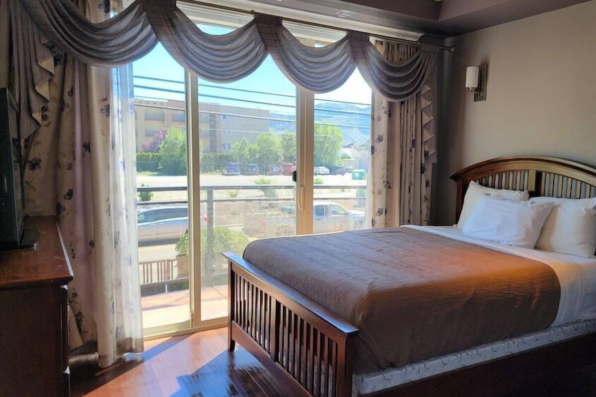 A bedroom with a large bed, a wooden headboard, a nightstand, a dresser, and a window with curtains.