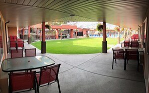 A covered patio area with tables and chairs, a grassy lawn, and a building with a red facade in the background.