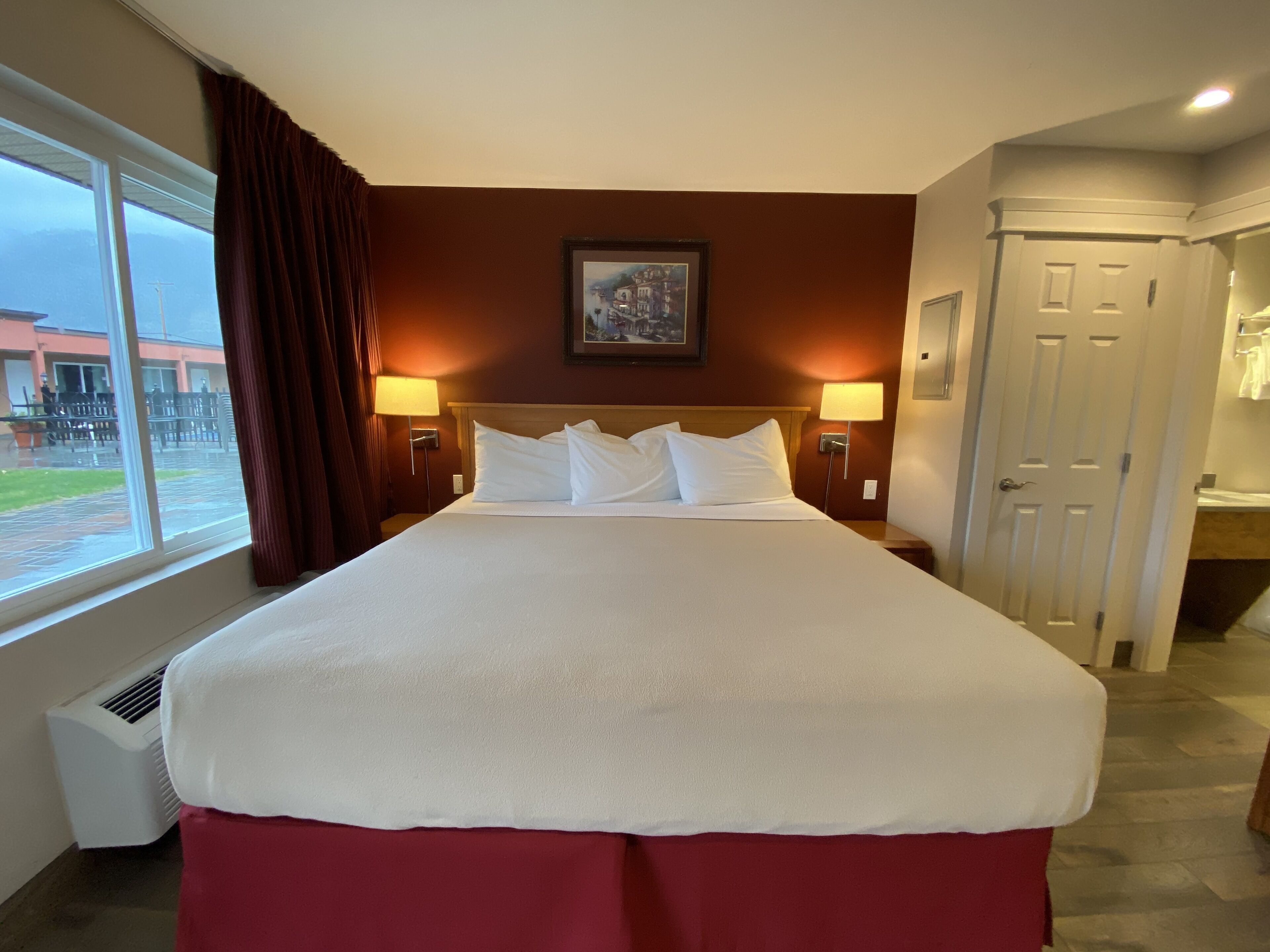 A hotel room with a large bed, two bedside lamps, a painting on the wall, and a door to the bathroom.