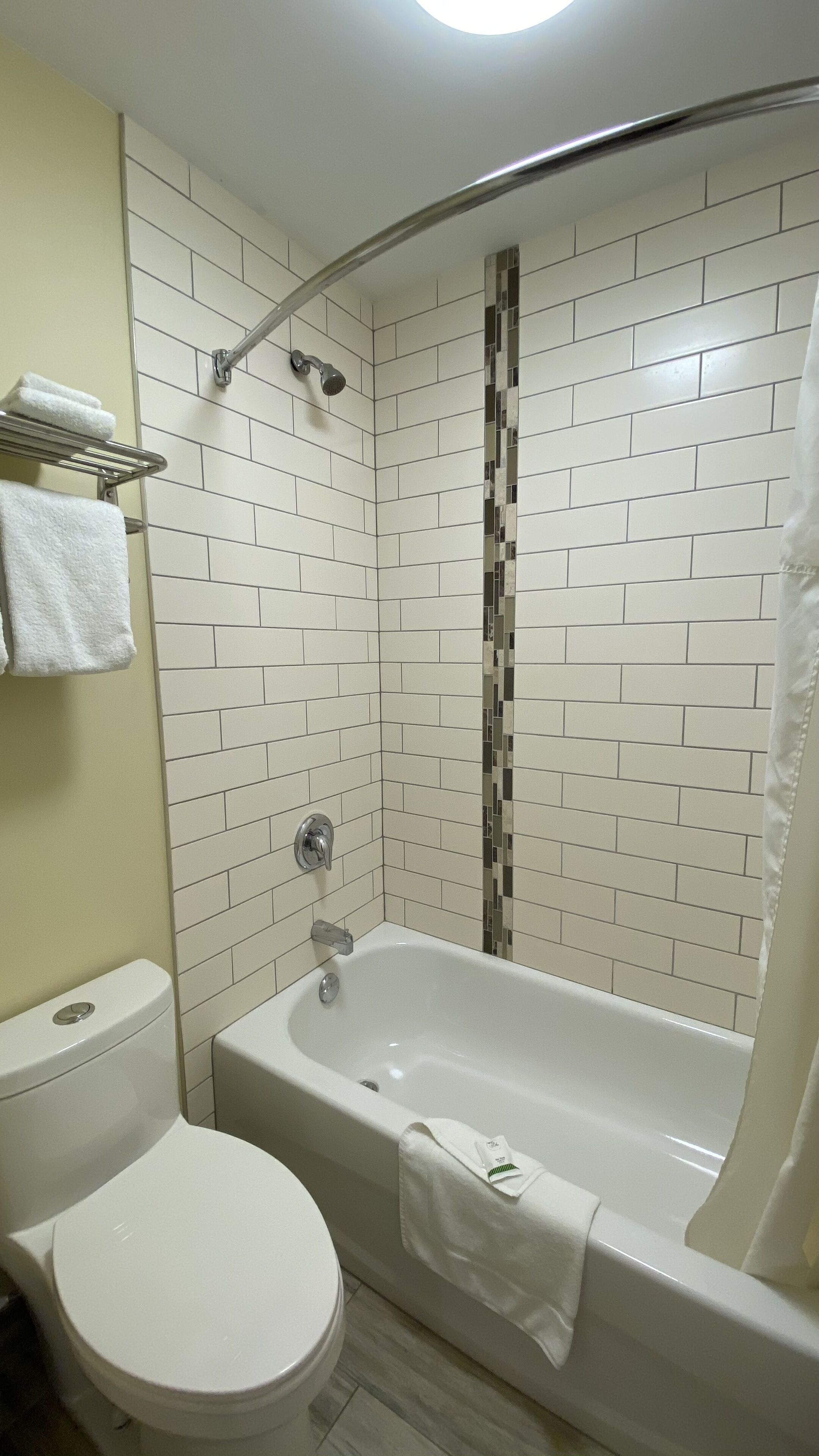 A bathroom with a white bathtub, a toilet, and a shower area with white tiles and a mosaic strip.
