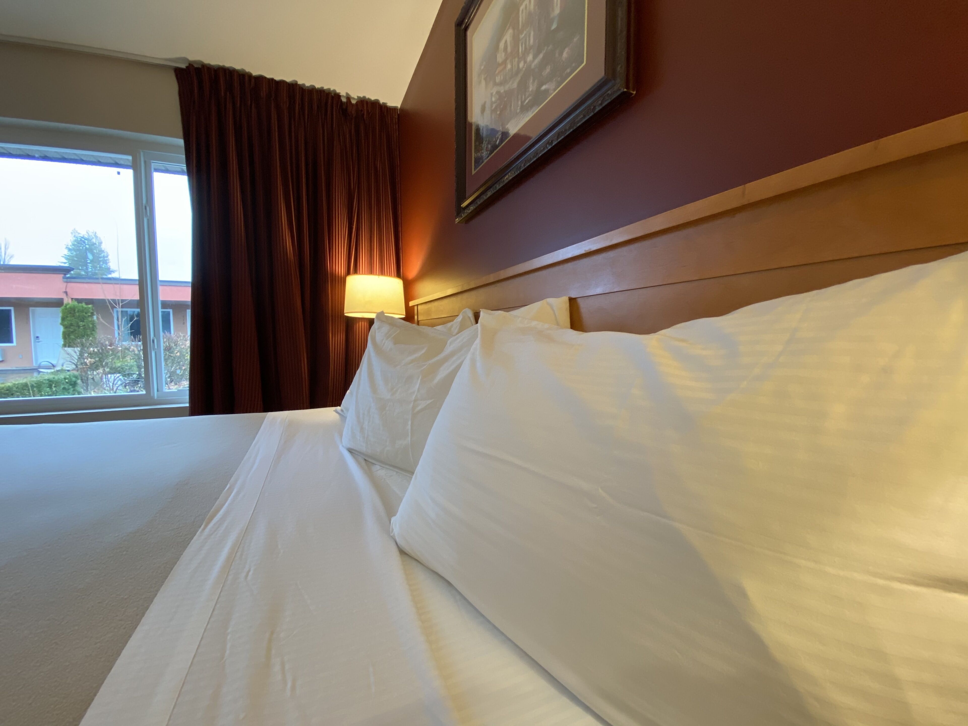 Quarto de hotel com uma cama grande, janela com cortinas, luminária fixada na parede e um quadro emoldurado.