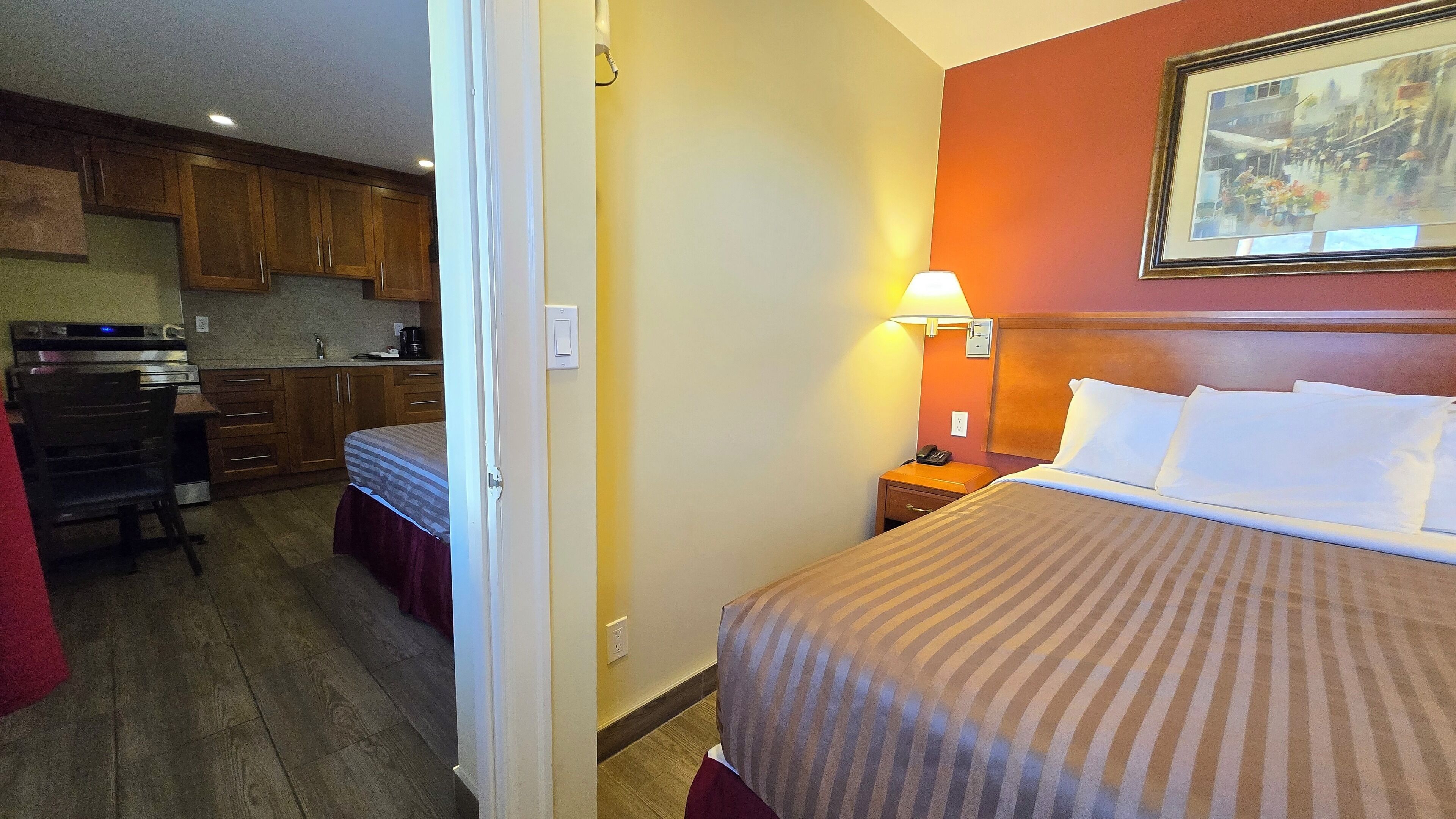 Una habitación de hotel con una cama, una mesita de noche con lámpara, un cuadro en la pared y una kitchenette en la habitación contigua.
