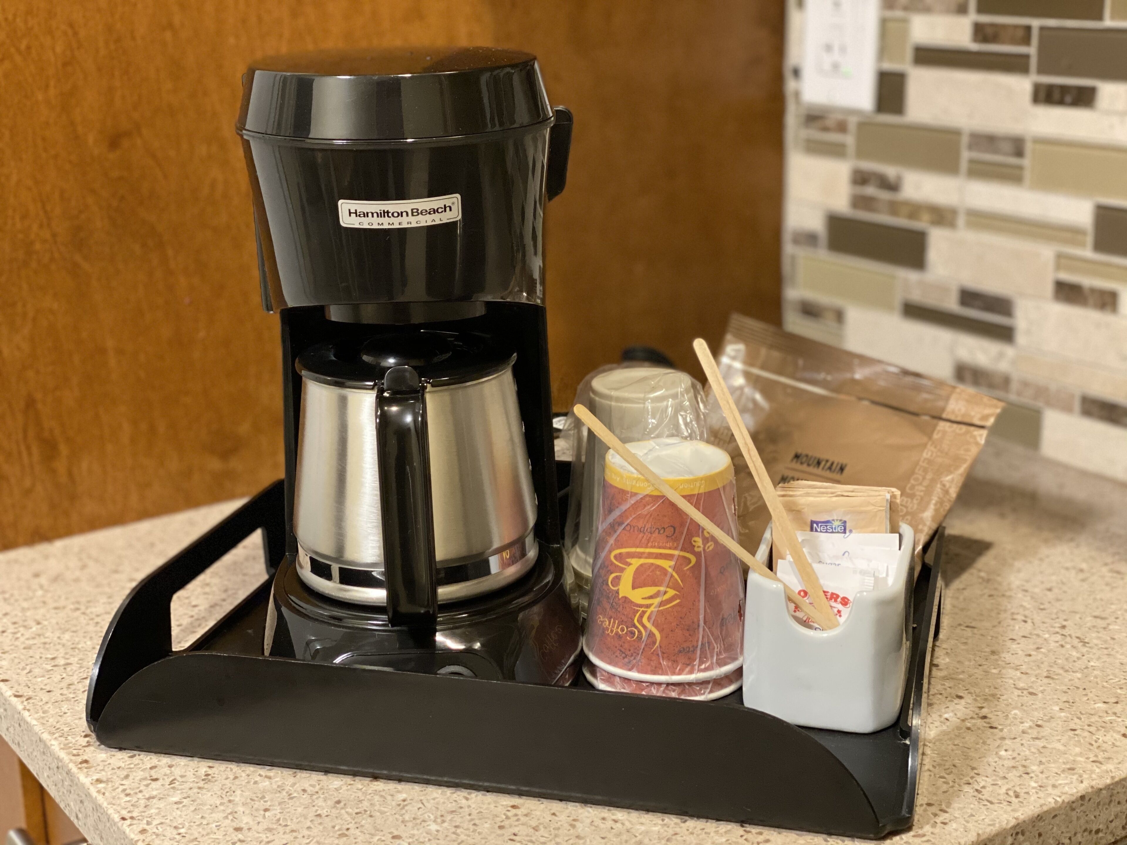 Une machine à café de marque « Hamilton Beach Commercial » est posée sur un comptoir, à côté d’un plateau, d’une tasse à café, d’une grande tasse et d’un paquet de dosettes de café.