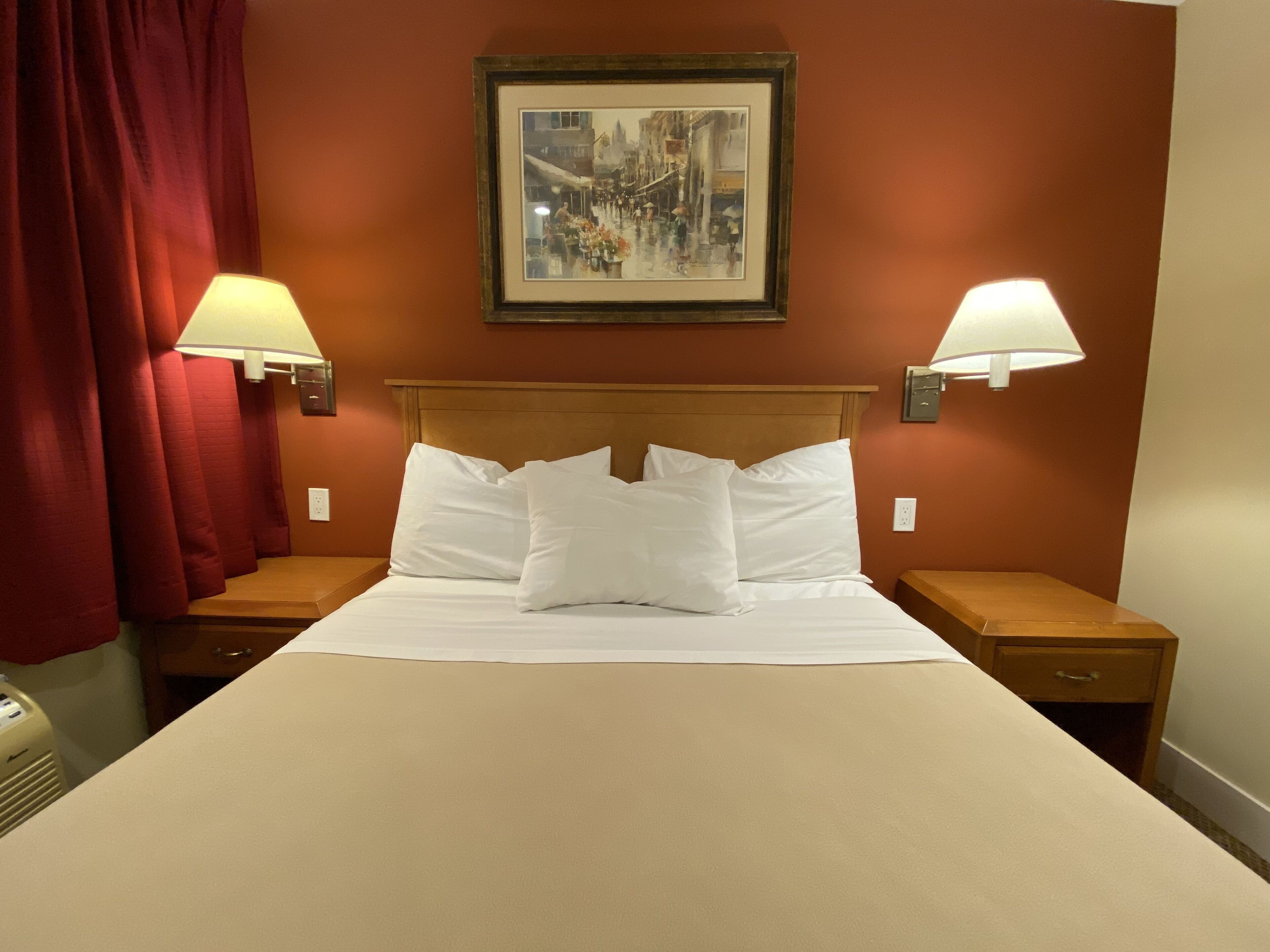 A hotel room with a large bed, two bedside lamps, a framed picture on the wall, and red curtains.