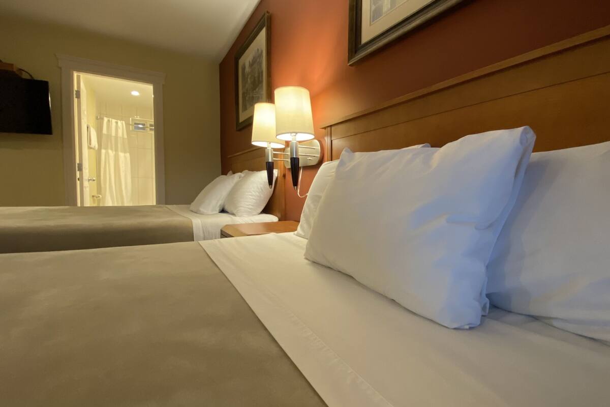 A hotel room with two beds, white linens, a wooden headboard, and a wall-mounted lamp.