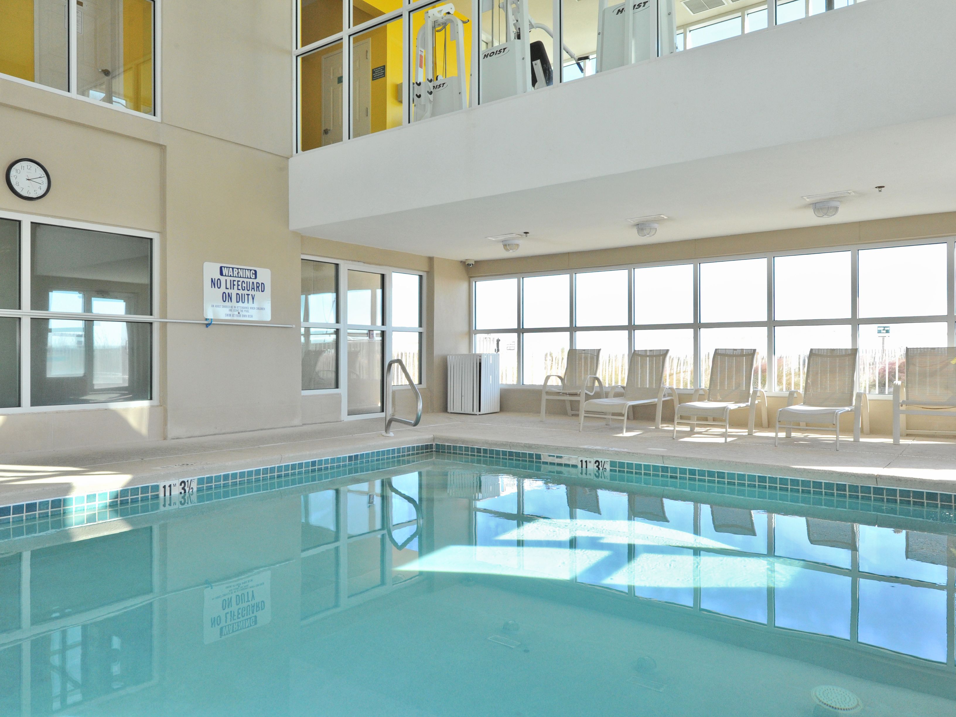 An indoor swimming pool with clear water, a clock on the wall, and a sign indicating no lifeguard on duty.