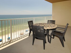 A balcony with a round table and four chairs, overlooking a beach with a few umbrellas and a distant ship.