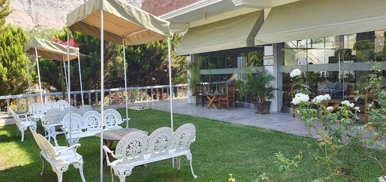 An outdoor patio with white metal chairs and tables, covered by beige canopies, and surrounded by lush greenery and potted plants.