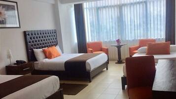 Een hotelkamer met een groot bed, een kleiner bed, een bureau en een stoel. Er is een raam met gordijnen en een lijst met een afbeelding aan de muur.