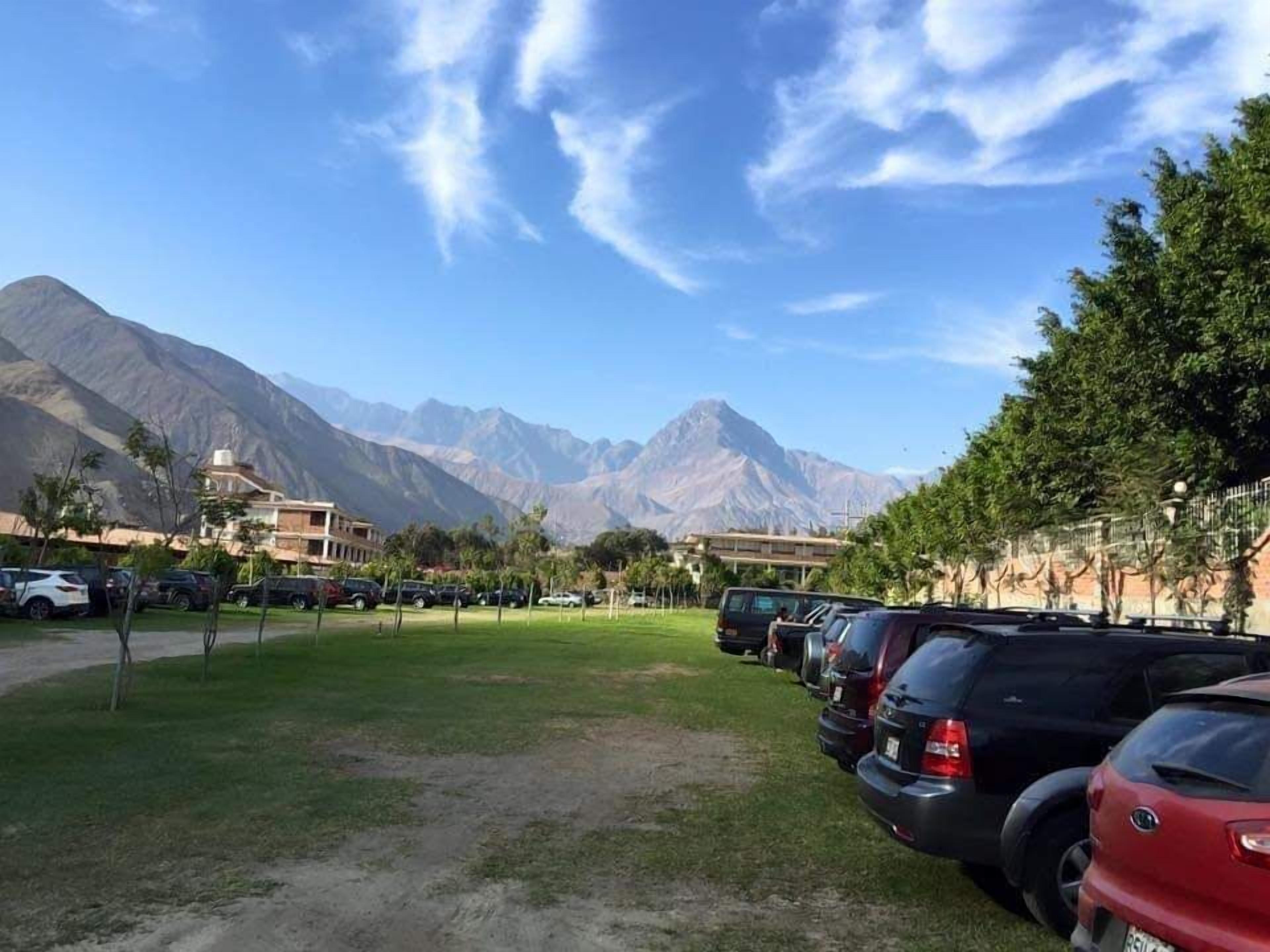Un estacionamiento con coches, una zona ajardinada y montañas al fondo.