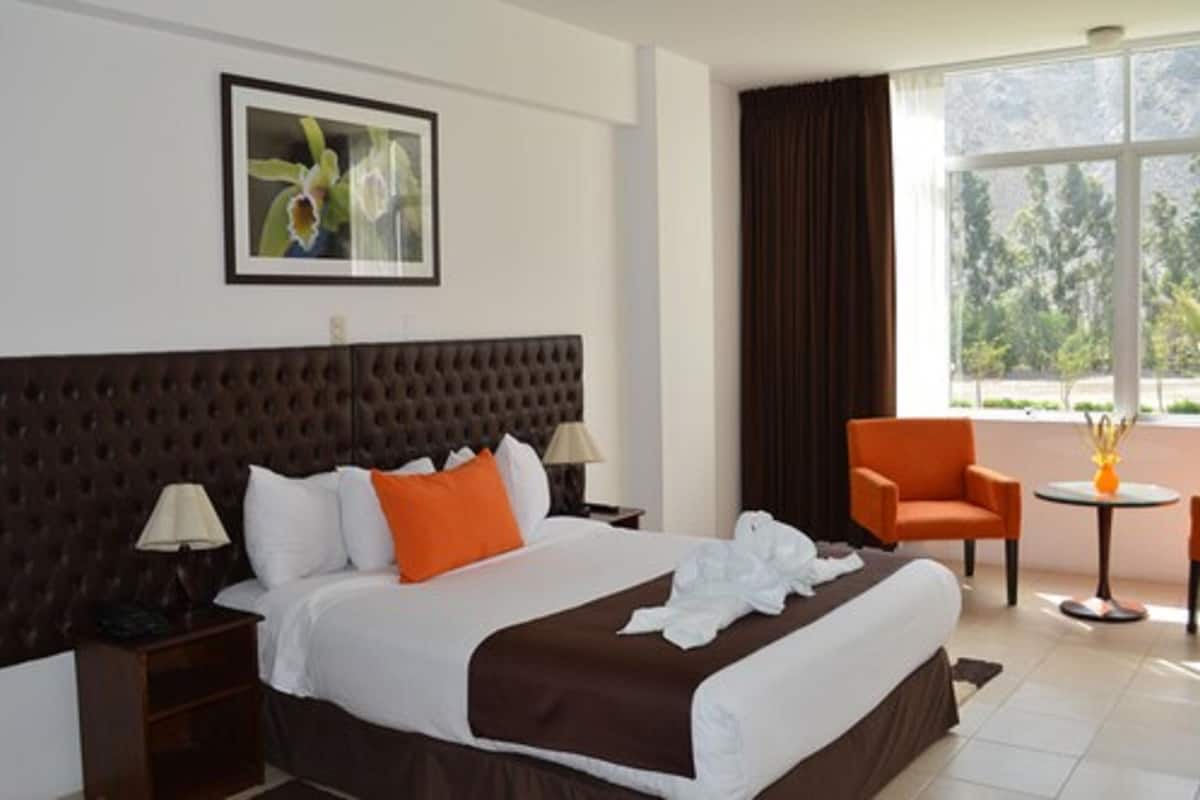 Ein Hotelzimmer mit einem großen Bett, einem Kopfteil, einem Nachttisch, einer Lampe, einem orangefarbenen Sessel, einem kleinen Tisch mit einer Vase und einem Fenster mit Blick auf Bäume.