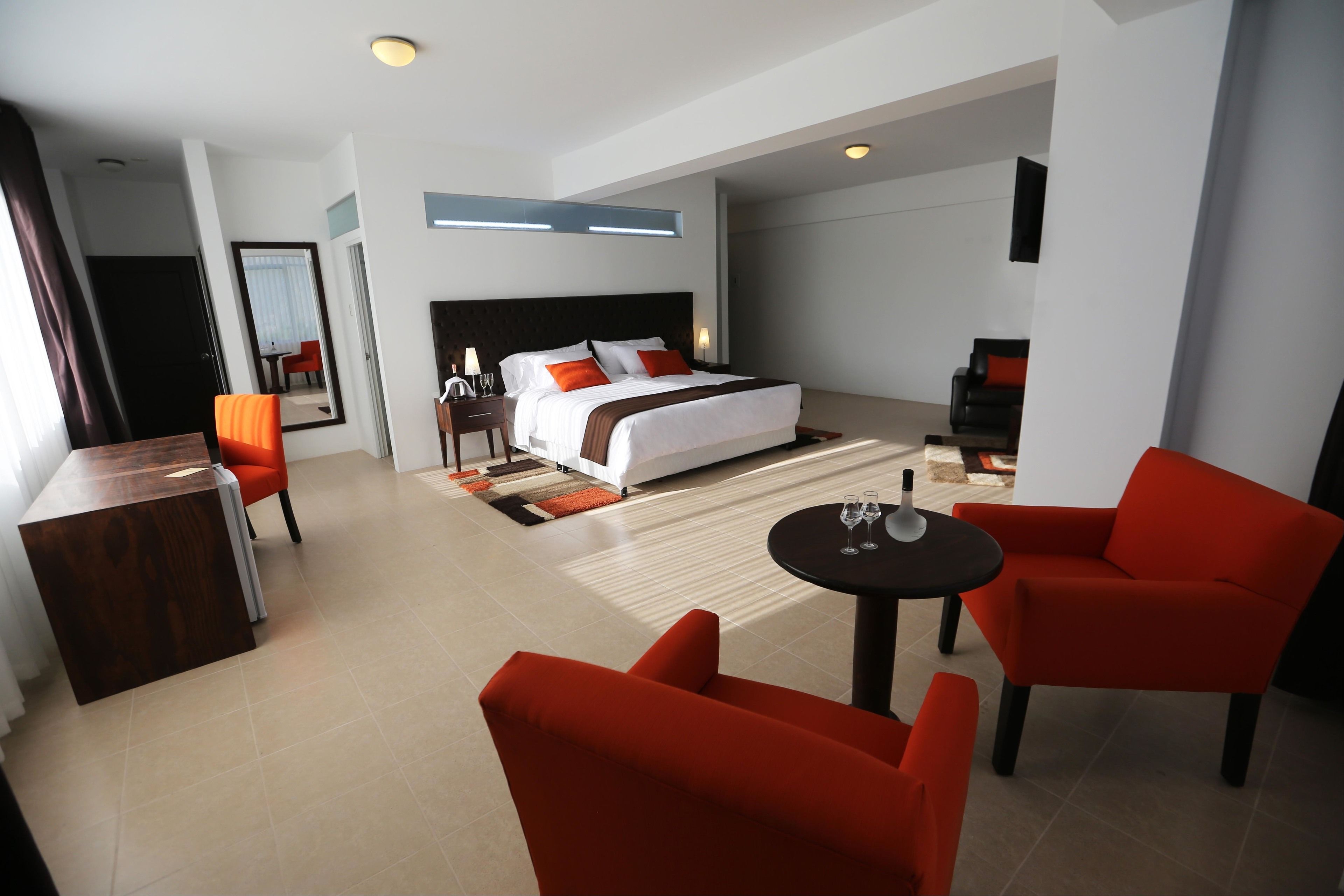 A modern hotel room with a large bed, a desk, two red chairs, and a small table with glasses.