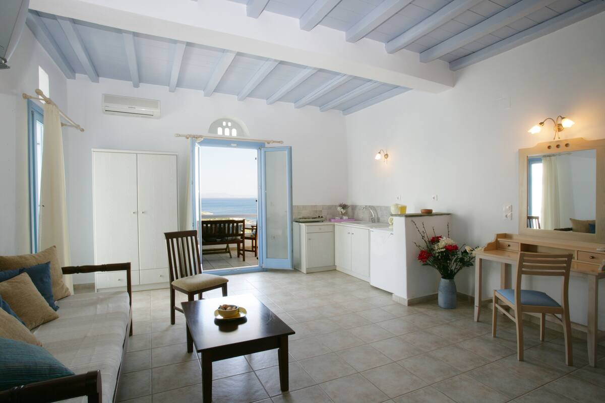 Amplia sala de estar con sofá, mesa de comedor y sillas. Se ve una cocina con un fregadero y alacenas. Hay un ventanal con vistas al exterior.