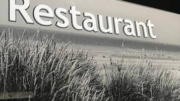 Ein Schild mit dem Wort 'Restaurant' in großen Buchstaben vor einer Kulisse aus hohem Gras und einer Strandlandschaft.